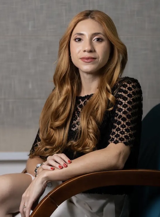 Natália Lima sentada em uma poltrona de madeira, com blusa preta de renda